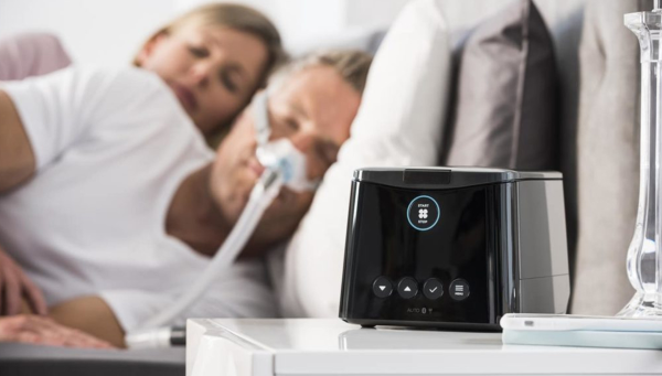 Fisher and Paykel SleepStyle CPAP machine on bedside table being used by a man using the brevida nasal mask in bed. 